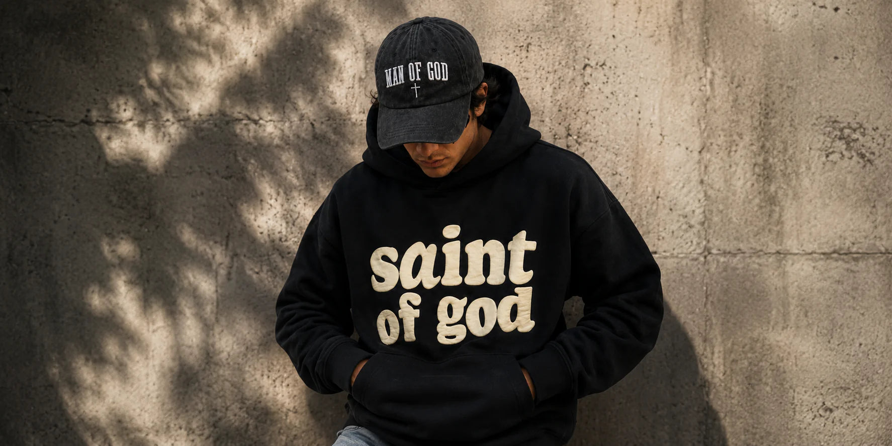 Person wearing a black hoodie with 'saint of god' text and a 'man of god' baseball cap by Soulful Faith against a textured wall.